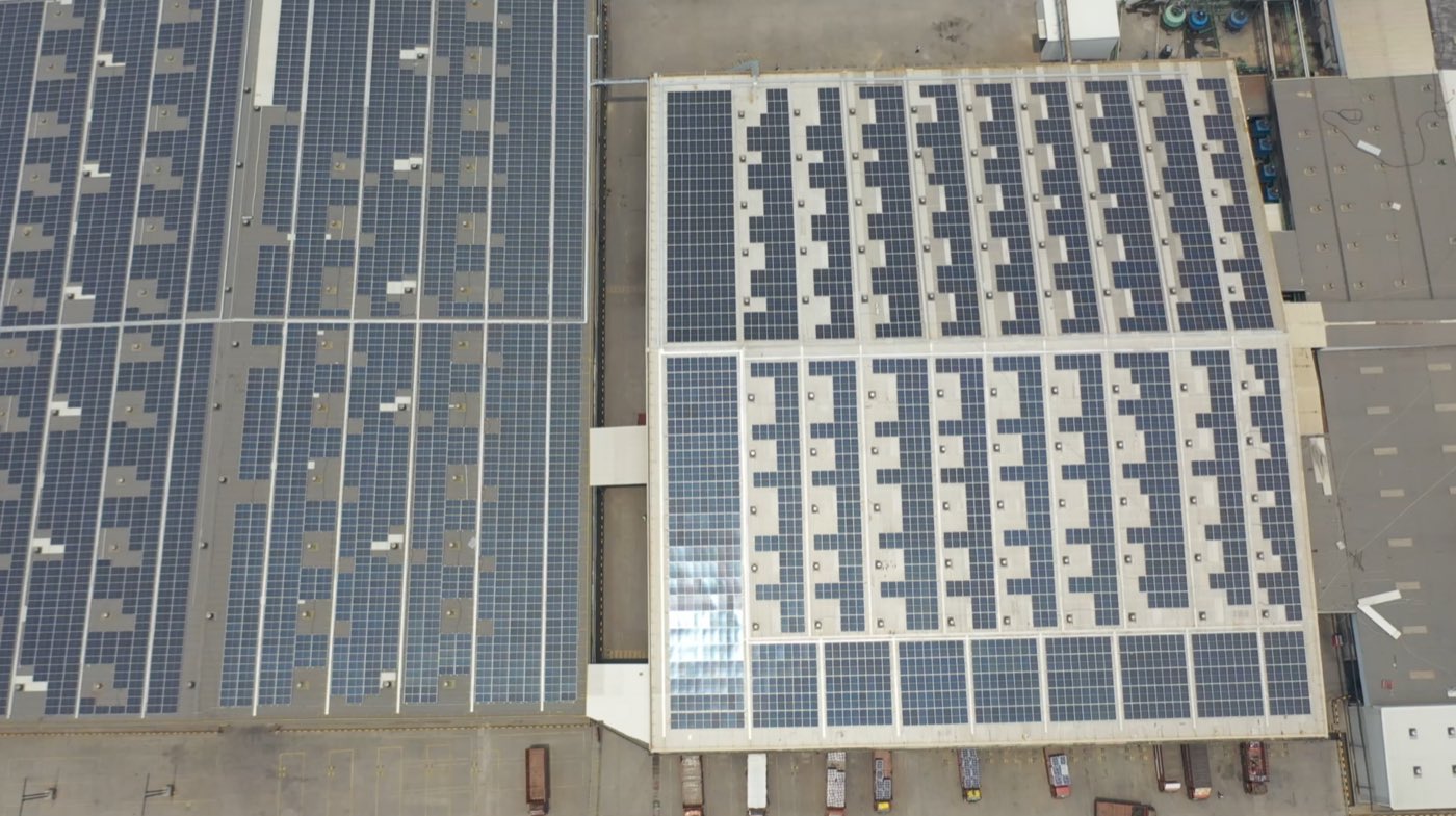 Top-down aerial view of the Dasna plant, two adjacent production hall roofs almost
                entirely covered with solar modules, with loading bays and delivery trailers below