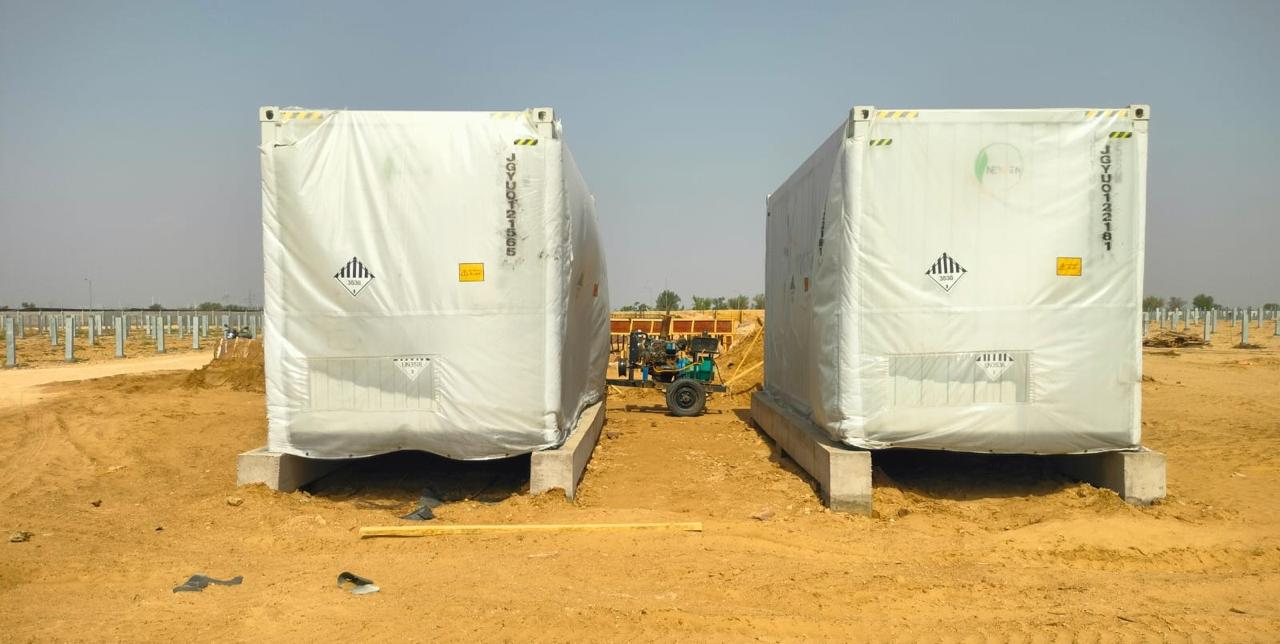Two wrapped battery containers standing on concrete plinths on site, with rows of mounting posts behind them