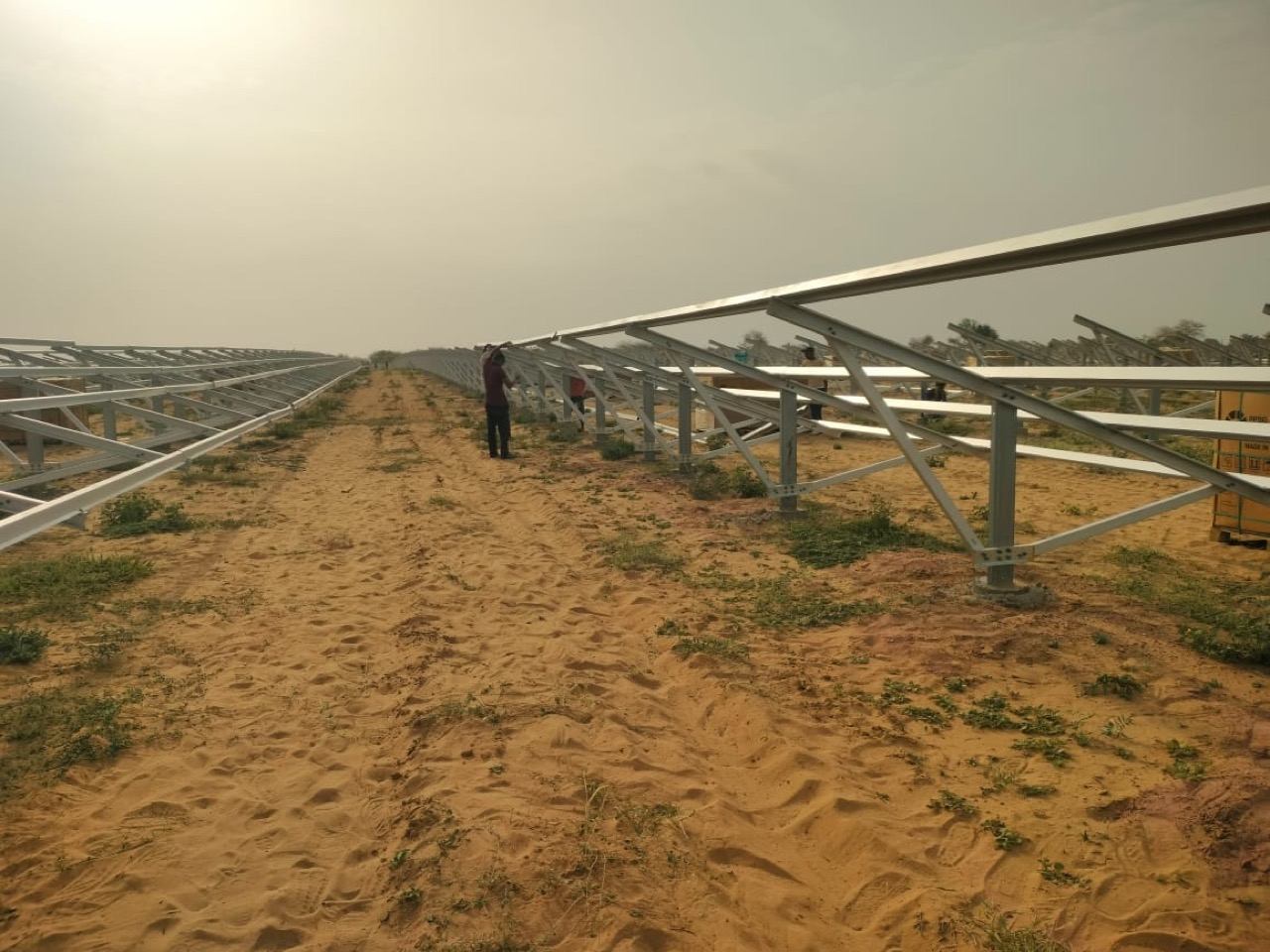 Workers erecting module mounting structures in a row across sandy desert ground, steel purlins running to the horizon